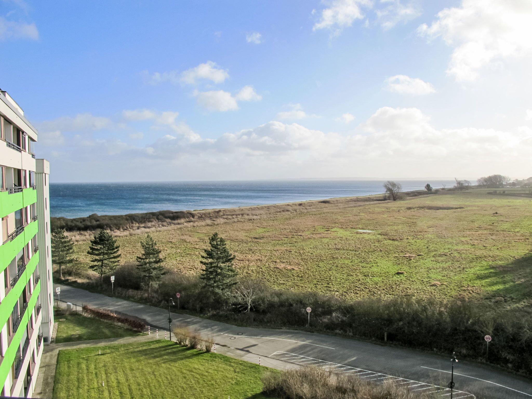 A view from a balcony overlooking a grassy field, a road, and the ocean.