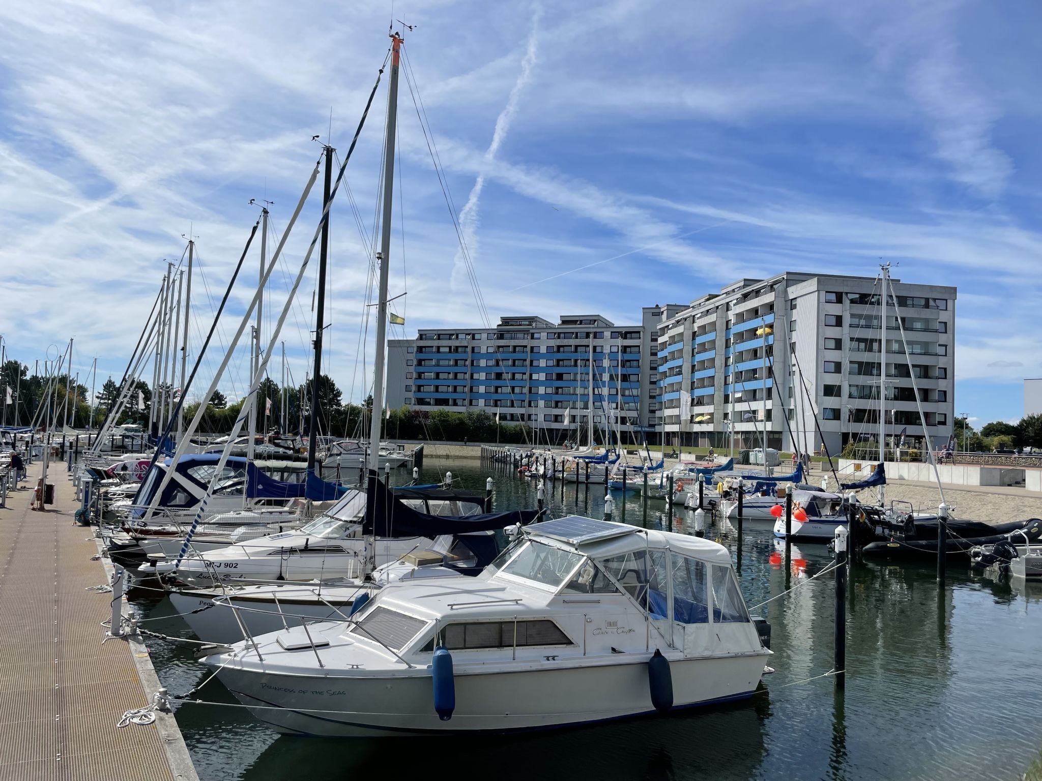 Un port de plaisance avec plusieurs bateaux amarrés, un bâtiment moderne en arrière-plan et un ciel dégagé.