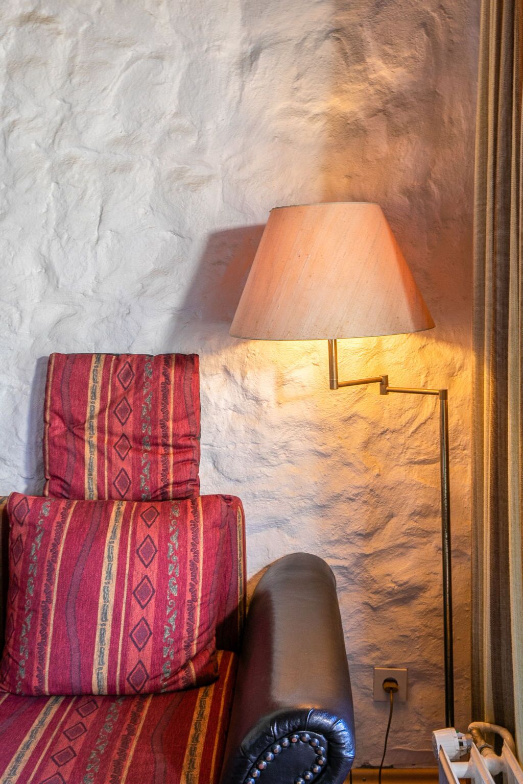 Ein Zimmer mit einer strukturierten weißen Wand, einem roten Kissen mit Muster, einem schwarzen Ledersofa und einer Stehlampe mit beiger Lampenschirm.