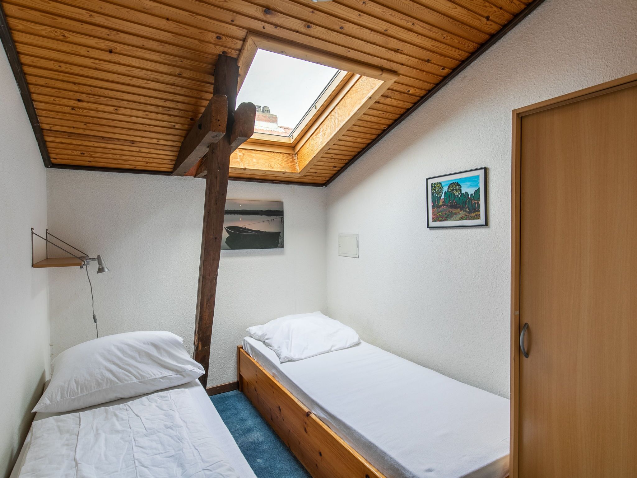 Une petite chambre accueillante, avec deux lits simples, une lucarne, un bureau avec une lampe et un tableau encadré au mur.