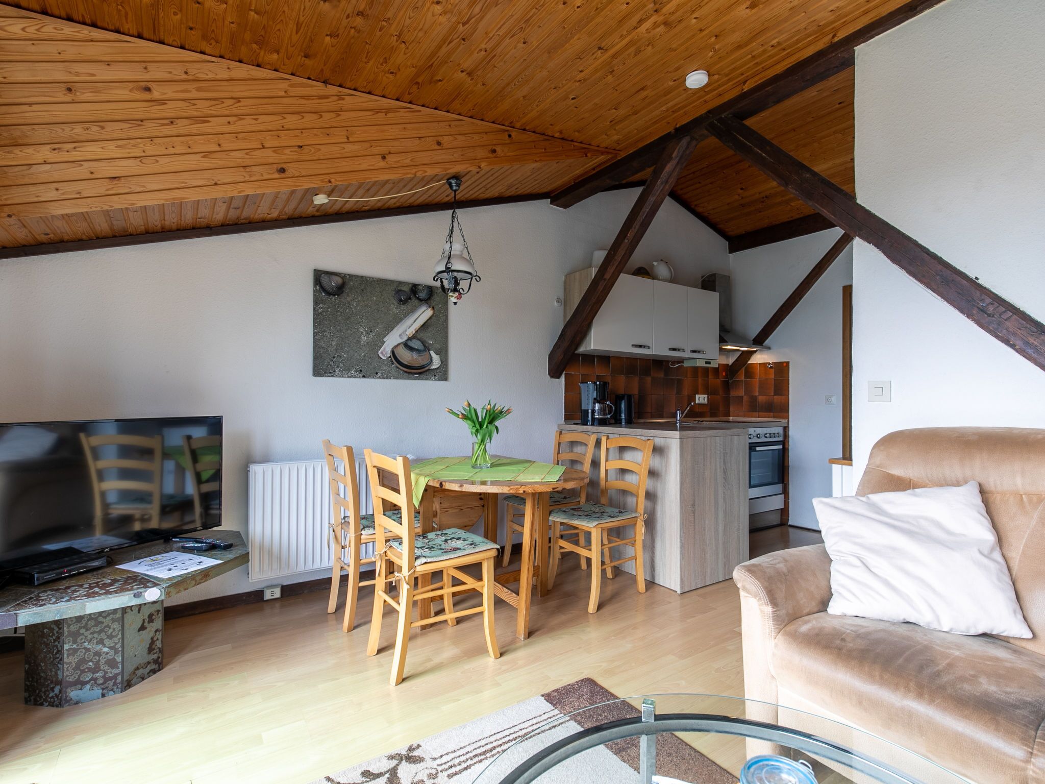 A cozy living room with a wooden ceiling, a dining area with a table and chairs, a sofa, and a kitchen in the background.