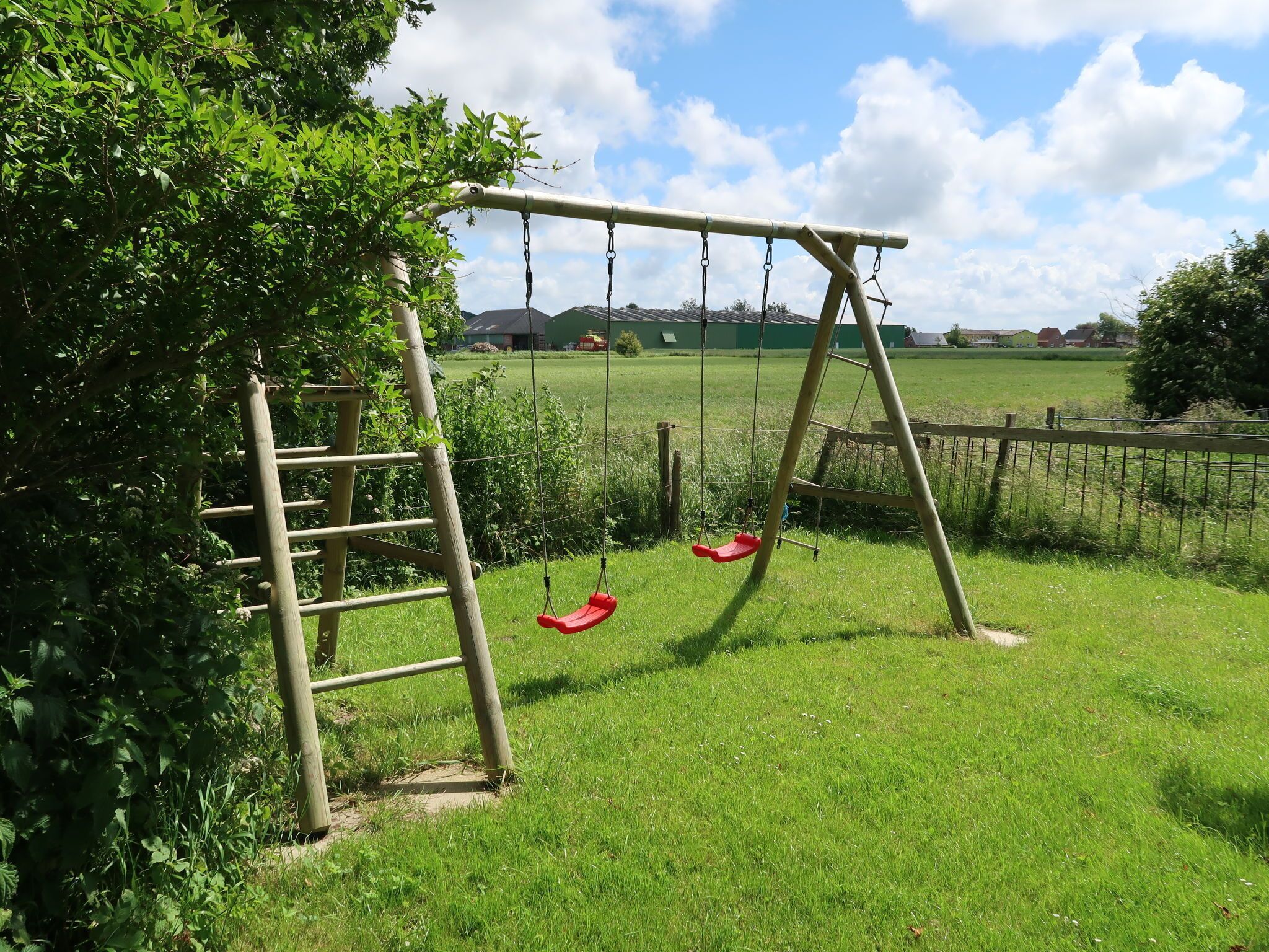 A wooden swing set in a grassy area with a ladder and a metal fence in the background.
