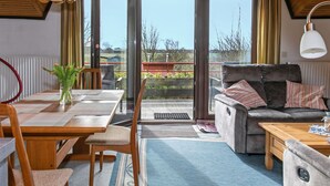 A cozy living room with a wooden ceiling, a sofa, a dining table with chairs, and a view through large glass doors to the outdoors.
