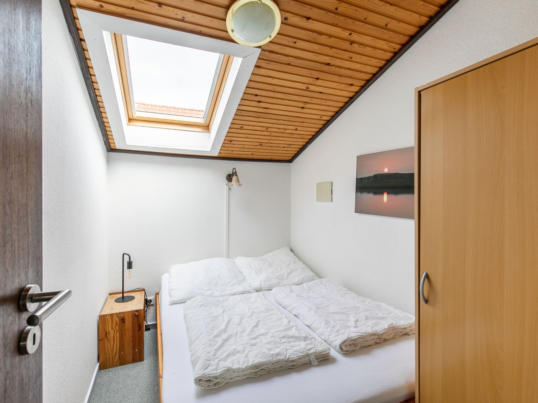 Un dormitorio pequeño con un tragaluz, una cama con ropa de cama blanca, una mesita de noche, una lámpara y una puerta de madera.