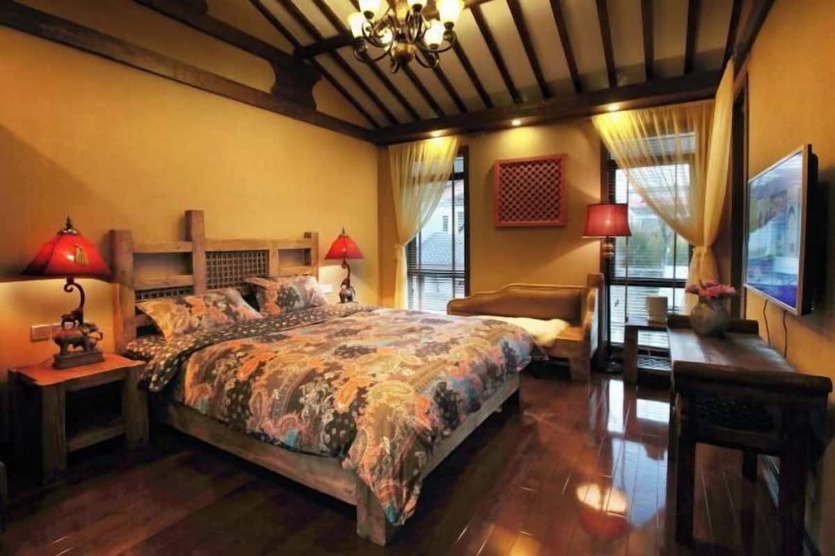 A bedroom with a large bed, wooden headboard, two bedside tables with lamps, a nightstand, a wooden bench, and a window with curtains.