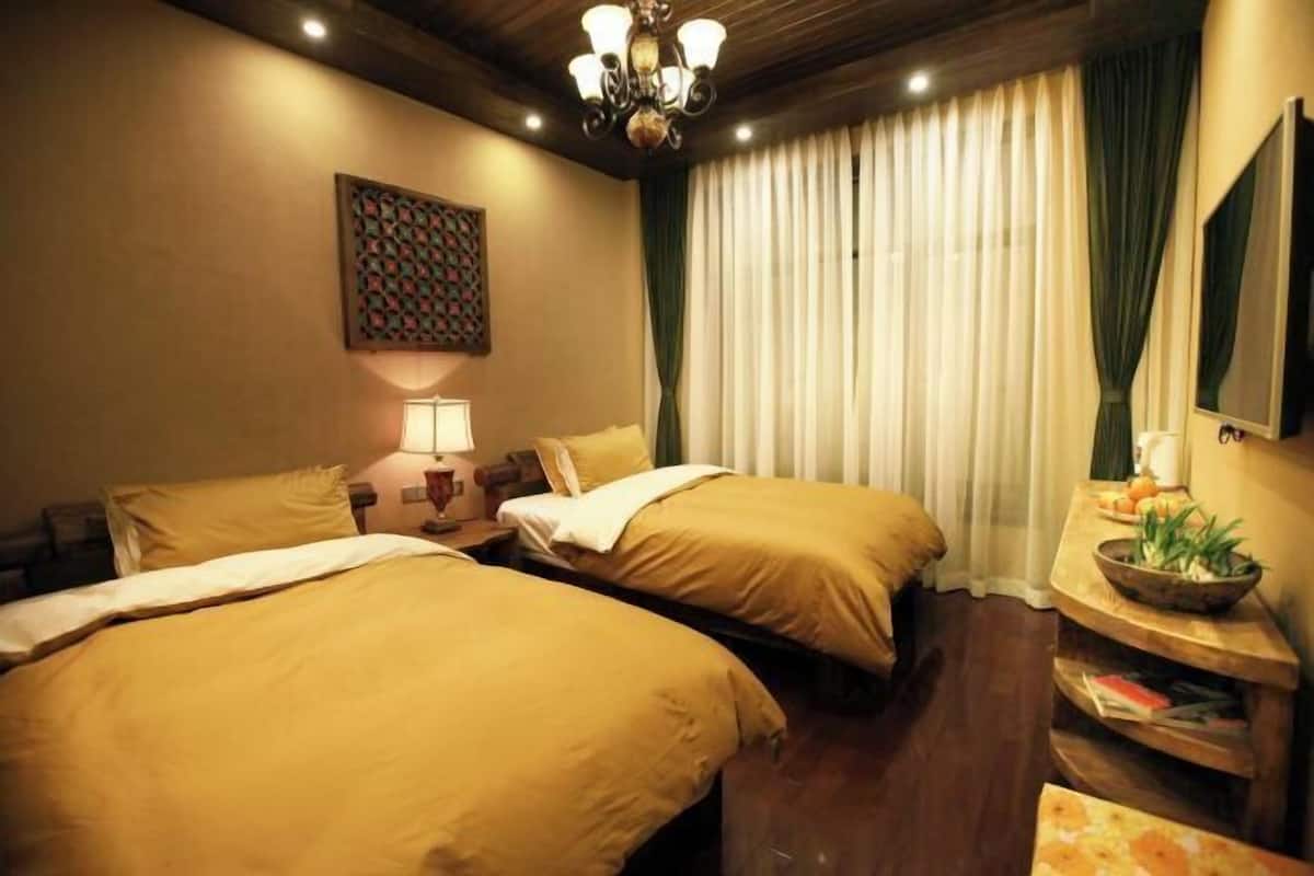 A hotel room with two beds, a chandelier, a painting, a window with curtains, and a small table with a bowl and books.