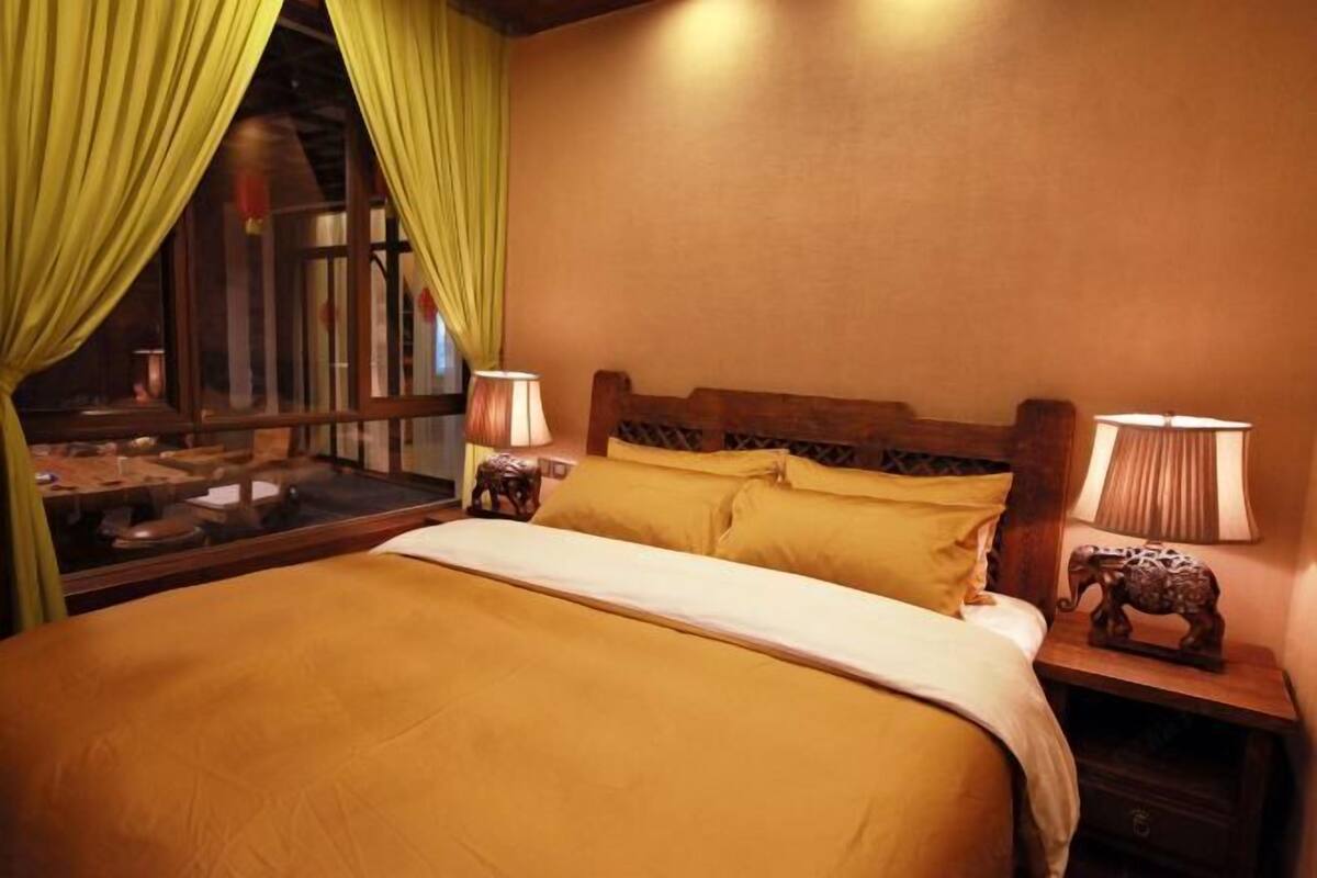 A bed with a wooden headboard, two bedside lamps, a nightstand with an elephant figurine, and a window with yellow curtains.