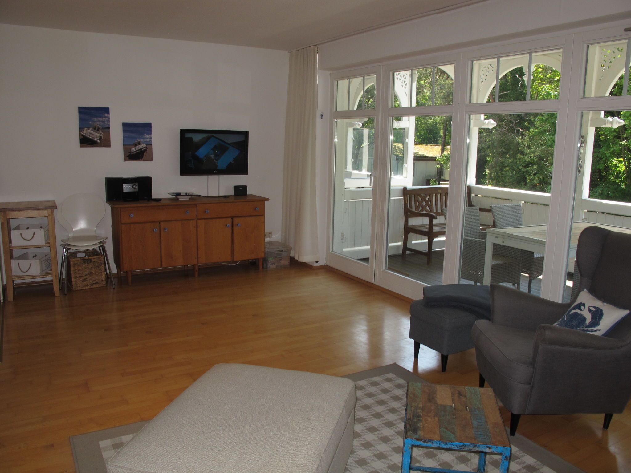Un salón espacioso con suelo de madera, un sofá gris, un aparador de madera, un televisor y ventanas amplias con vistas al exterior.