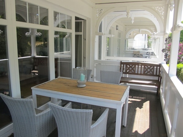 A covered outdoor area with a table and chairs, large windows, and a view of the garden.