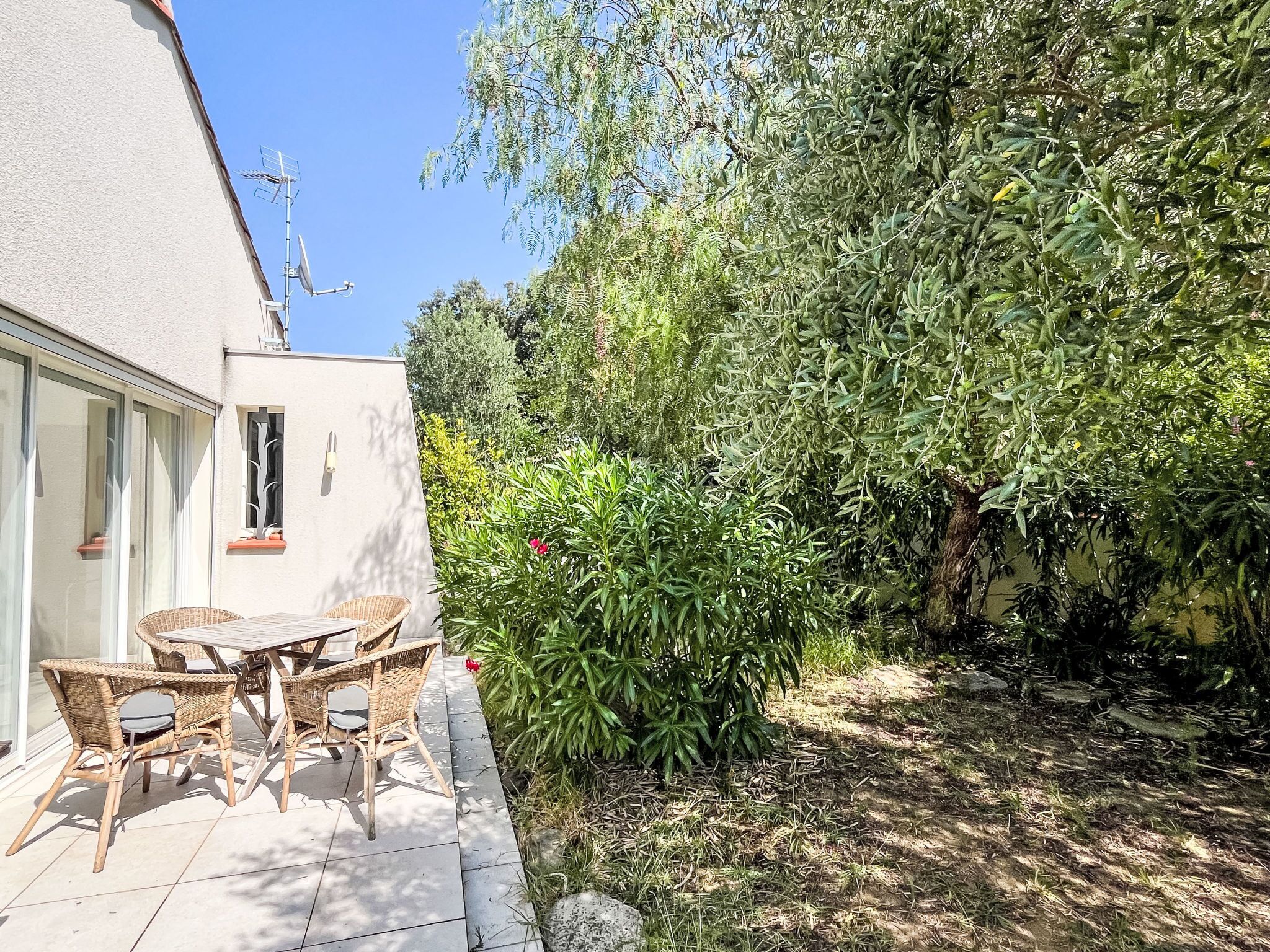 Une terrasse dotée de meubles en osier et offrant une vue sur un jardin où poussent différentes plantes et un arbre.