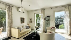 Ein modernes Wohnzimmer mit einem beigefarbenen Sofa, einem weißen Sessel und einem Glastisch. Zwei Fenster mit Vorhängen, eine Deckenleuchte und eine Vitrine mit Dekorationselementen sind ebenfalls zu sehen.