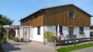 A two-story building with a wooden facade and a white lower section. There is a covered outdoor seating area with a bench and potted plants. - Osterende by Interhome (Otterndorf)