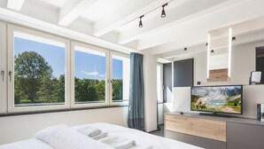 Ein modernes Hotelzimmer mit einem großen Bett, einem Flachbildfernseher und Blick auf Bäume durch die Fenster.