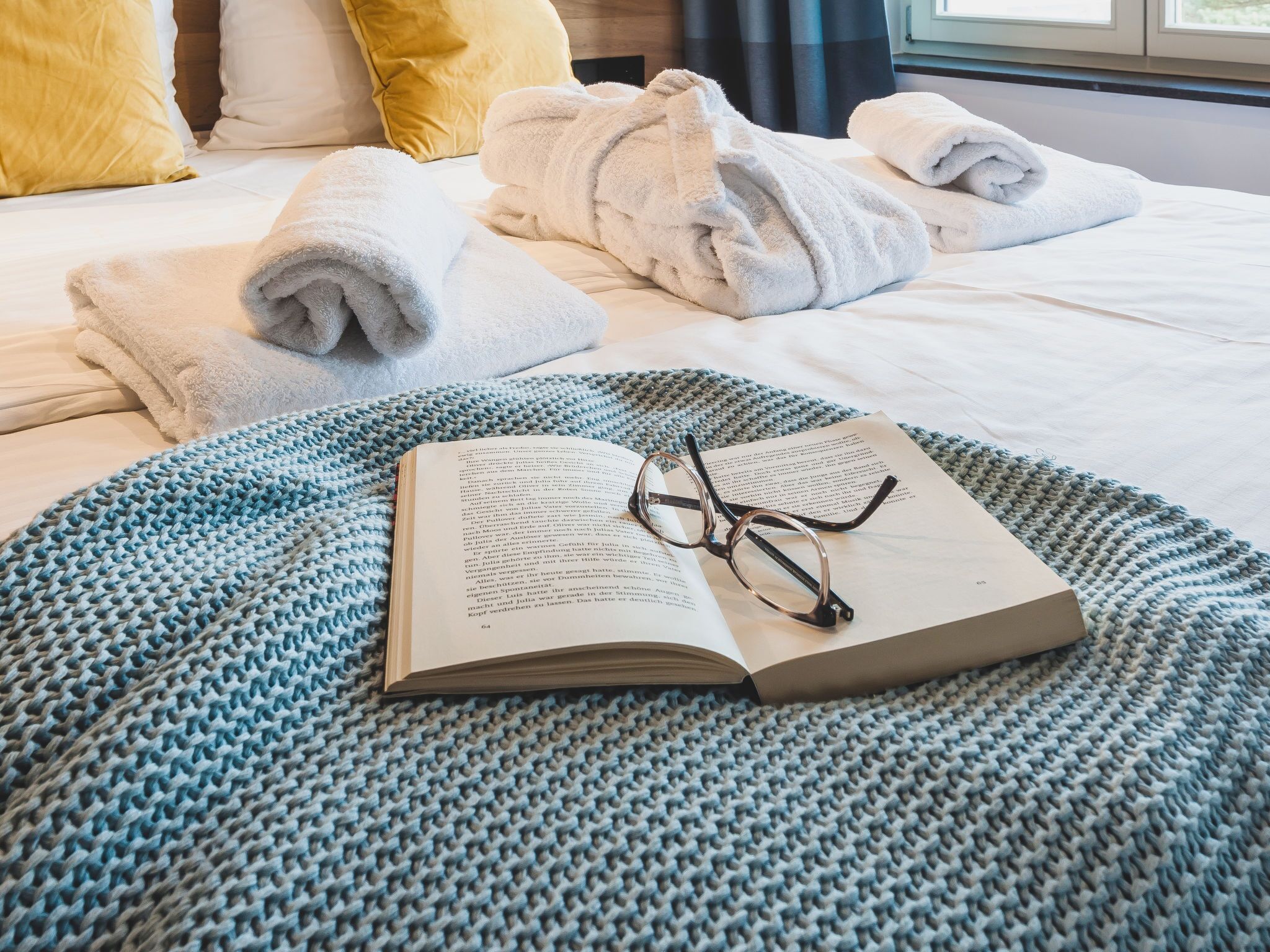 Un lit bien fait, avec des serviettes blanches pliées en rouleau, une couverture bleue tricotée et un livre sur lequel reposent des lunettes.