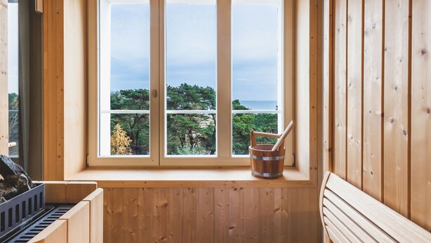 A wooden sauna room with a bench, a bucket with a stick, and a view of trees and water through a window. - Mariandl am Meer by Interhome (Ostseebad Binz)