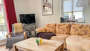 A living room with a wooden coffee table, a grey sofa, a red curtain, a TV, and a bookshelf with books and a wine bottle. - Boddenzauber by Interhome (Breege)