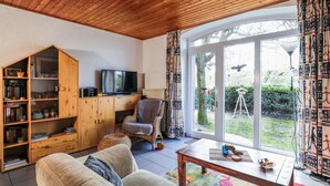A cozy living room with a wooden ceiling, a television on a wooden cabinet, a wicker chair, and a large window with curtains. - von Dollen by Interhome (Wingst)