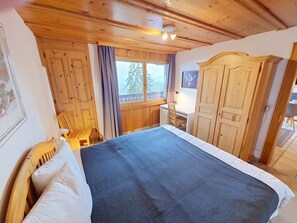 Ett sovrum i en stuga med träpanel, inrett med en säng, en stol, en byrå och ett fönster med utsikt över träd.
