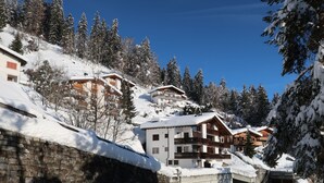 A snow-covered mountain village with chalet-style buildings, a stone wall, and a winding road.