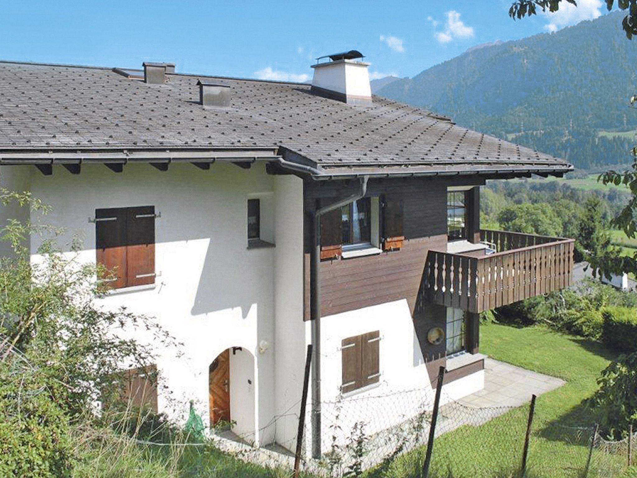Een tweelaags huis met een balkon, een houten terras en uitzicht op een bergachtig landschap.