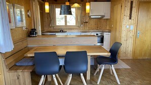 A wooden interior with a wooden table, two blue chairs, a kitchen area with cabinets and a sink, and pendant lights. - Chalet Soldanella (BEL205) by Interhome (Bellwald)