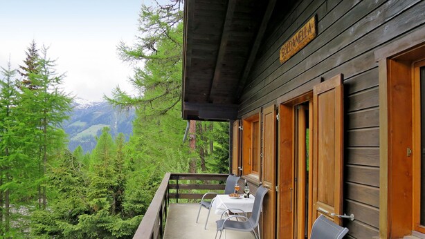 A wooden cabin with a balcony, furnished with tables and chairs, overlooking a forested mountain area. - Chalet Soldanella (BEL205) by Interhome (Bellwald)