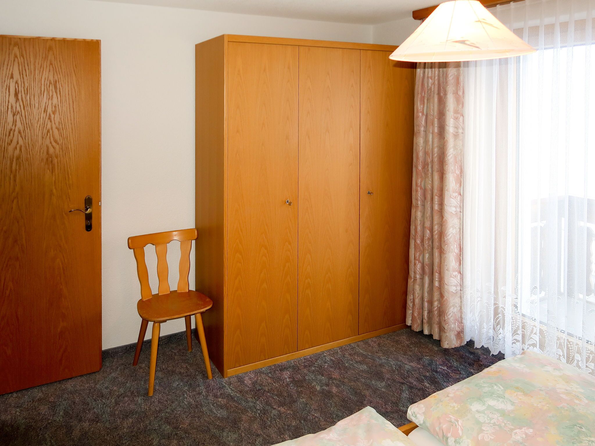 Quarto com guarda-roupa de madeira, cadeira de madeira, cama com roupa de cama floral e janela com cortinas leves.