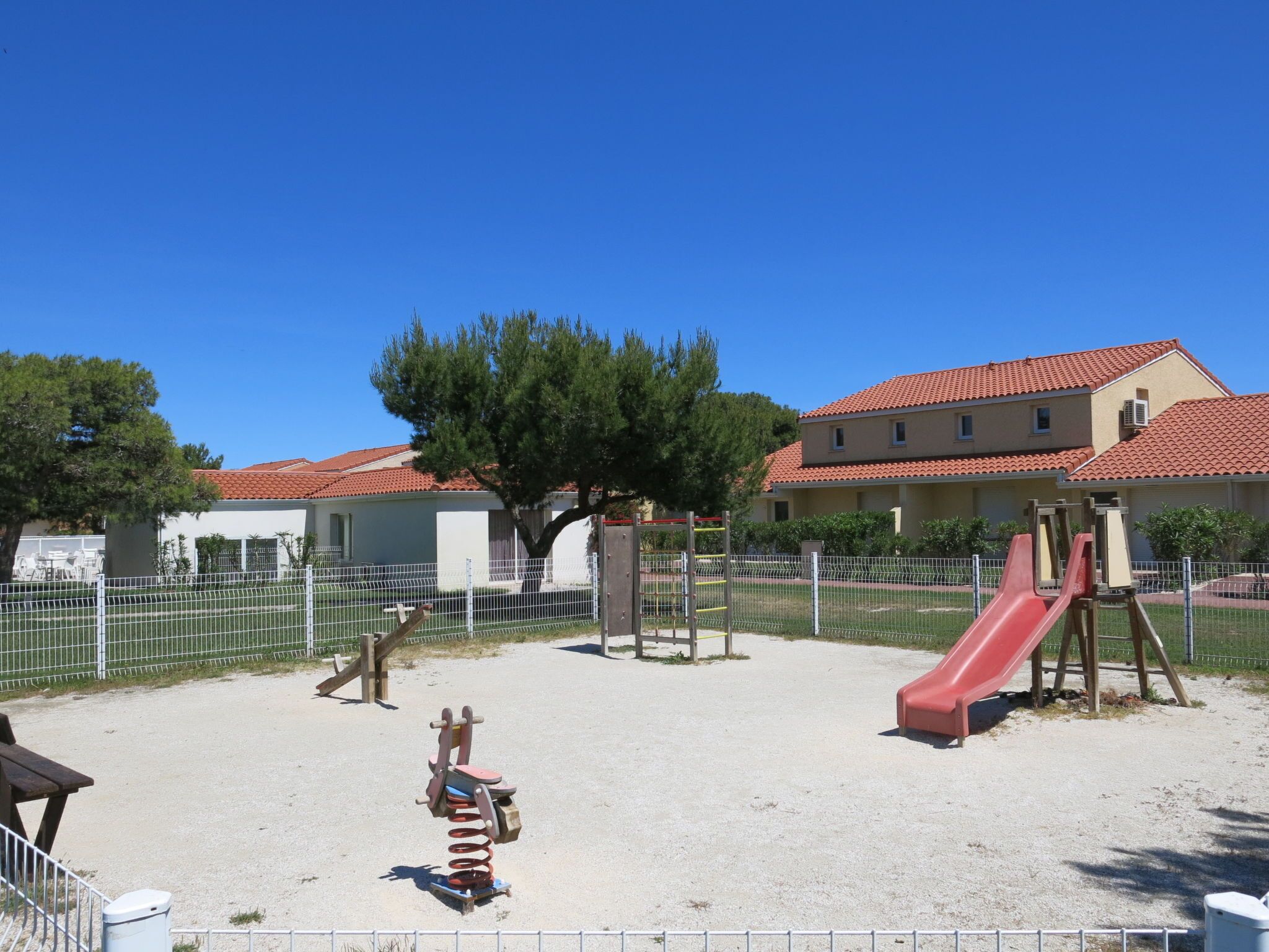 Ein Spielplatz mit Rutsche und Schaukeln, umgeben von einem Zaun und Bäumen.