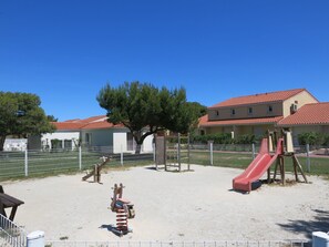 Un parque infantil con tobogán y columpios, rodeado por una valla y árboles.