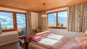 Une chambre confortable avec un plafond en bois, un lit recouvert d’une literie à motifs floraux, un radiateur et de grandes fenêtres offrant une vue sur les montagnes.