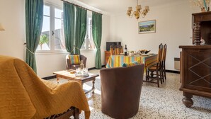 A cozy living room with a sofa, a dining table set for four, and a large window with green curtains. - La Vigneronne by Interhome (Fourques)