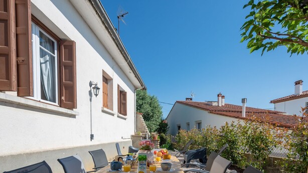 A dining area with a table set for breakfast, outdoor chairs, and a view of a white building with brown shutters. - La Vigneronne by Interhome (Fourques)