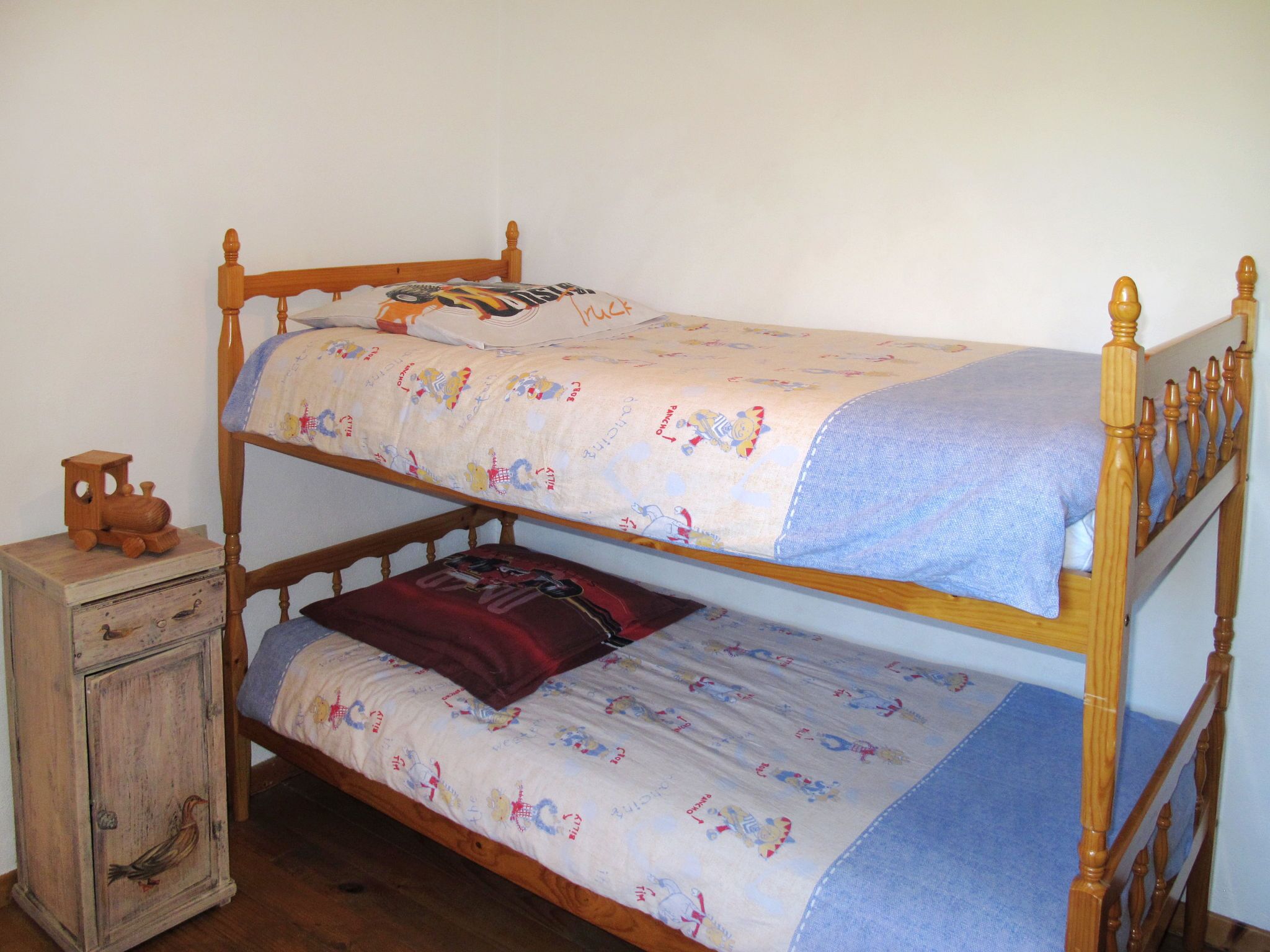 A bunk bed with a wooden frame and a lower bunk with a pillow. There is a small wooden chest of drawers with a bird design on the front.