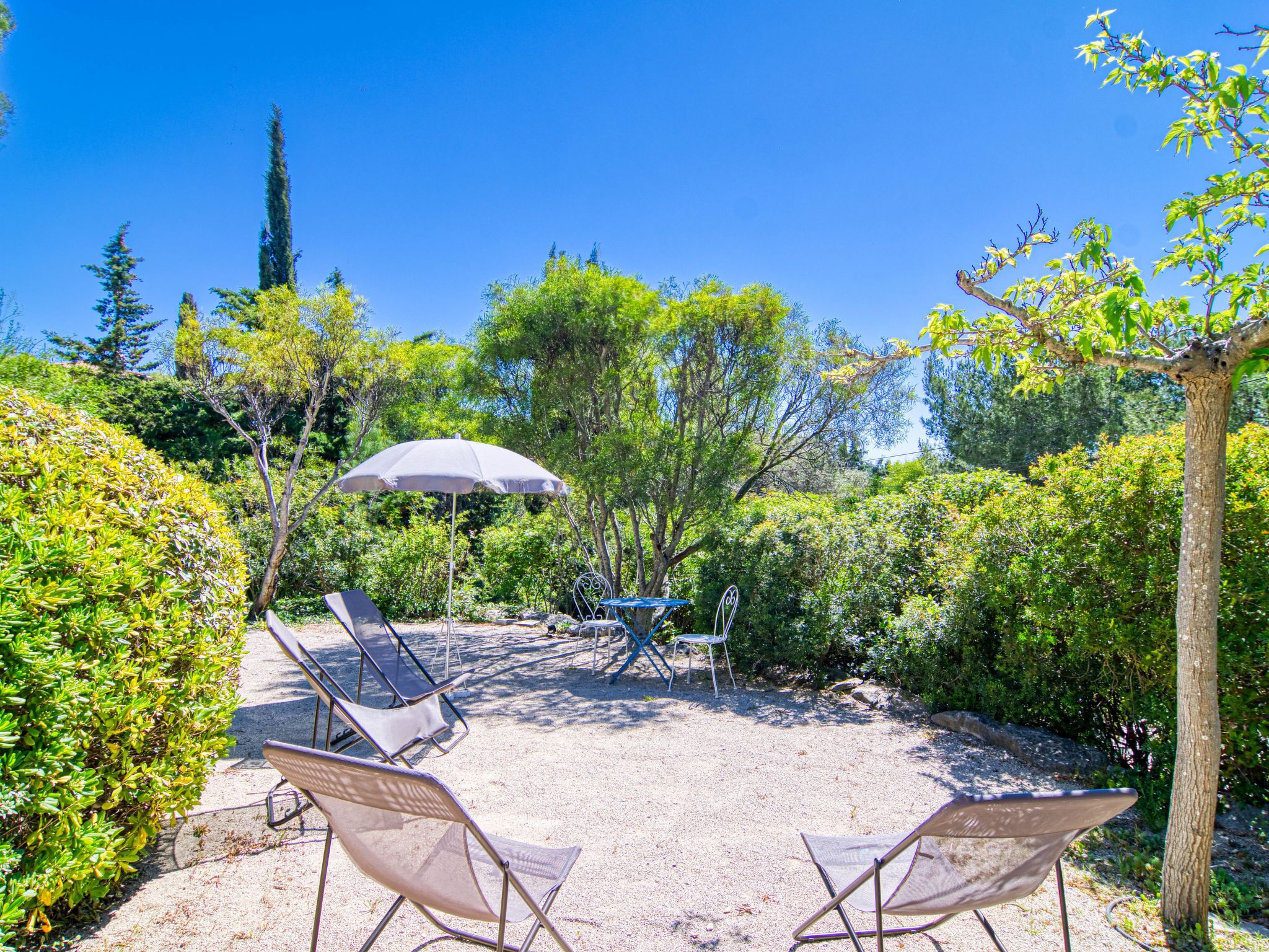An outdoor seating area with two lounge chairs and a table under a large umbrella, surrounded by lush greenery and trees.
