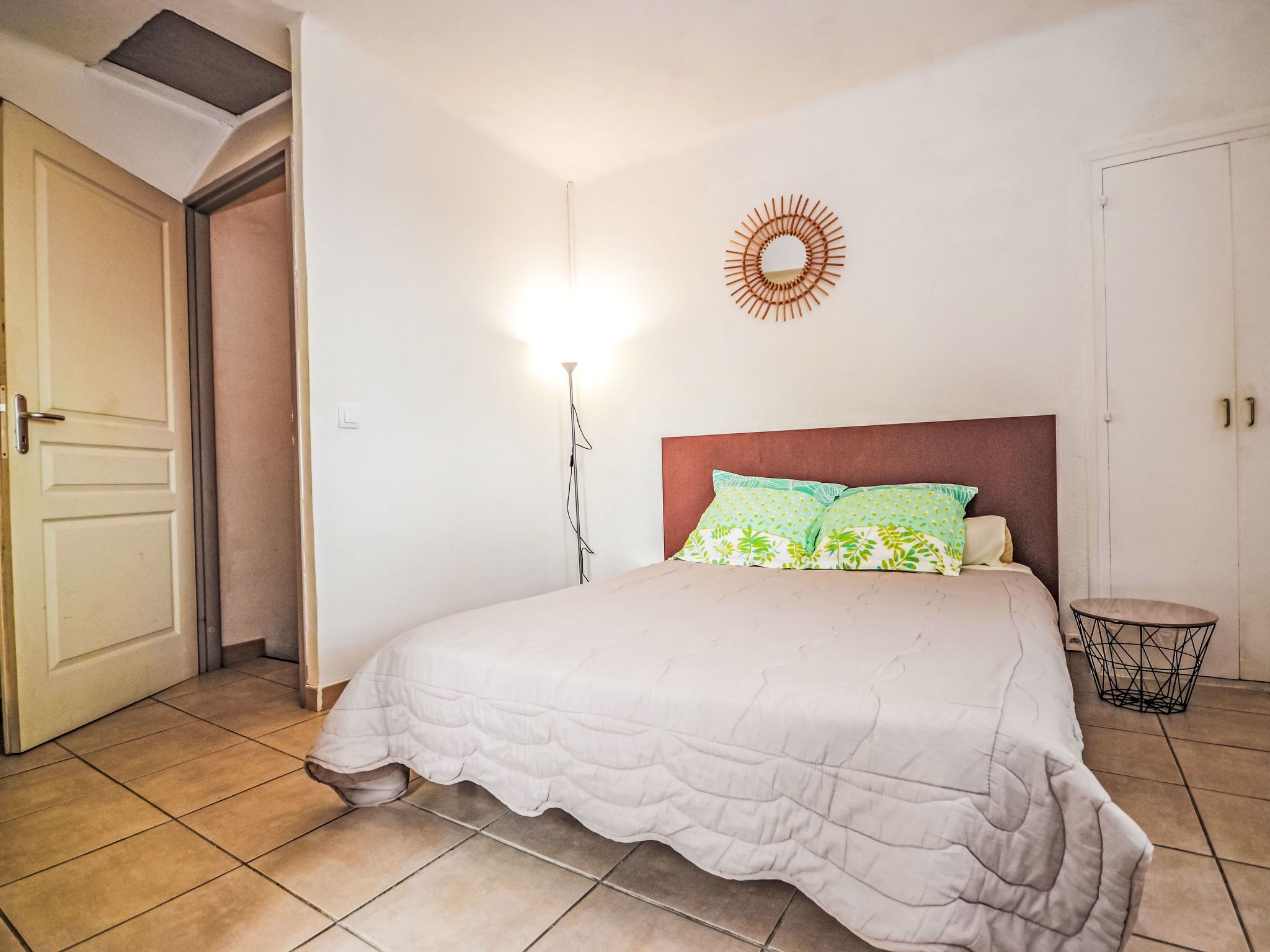Une chambre à coucher avec un lit, une lampe de chevet, un miroir rond accroché au mur et une porte.