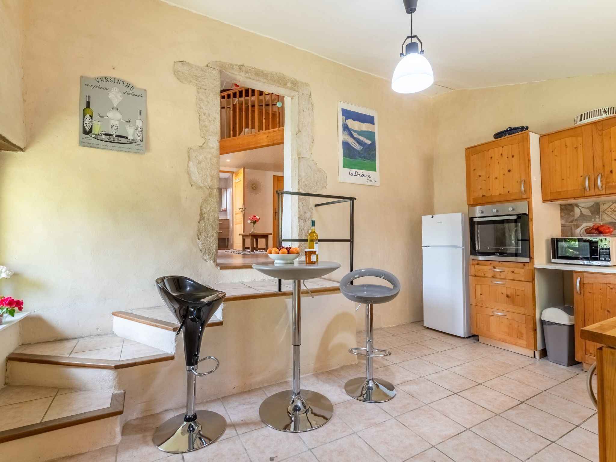 A kitchen with wooden cabinets, a white refrigerator, an oven, and a microwave. There are two bar stools and a small table with a bottle and oranges.