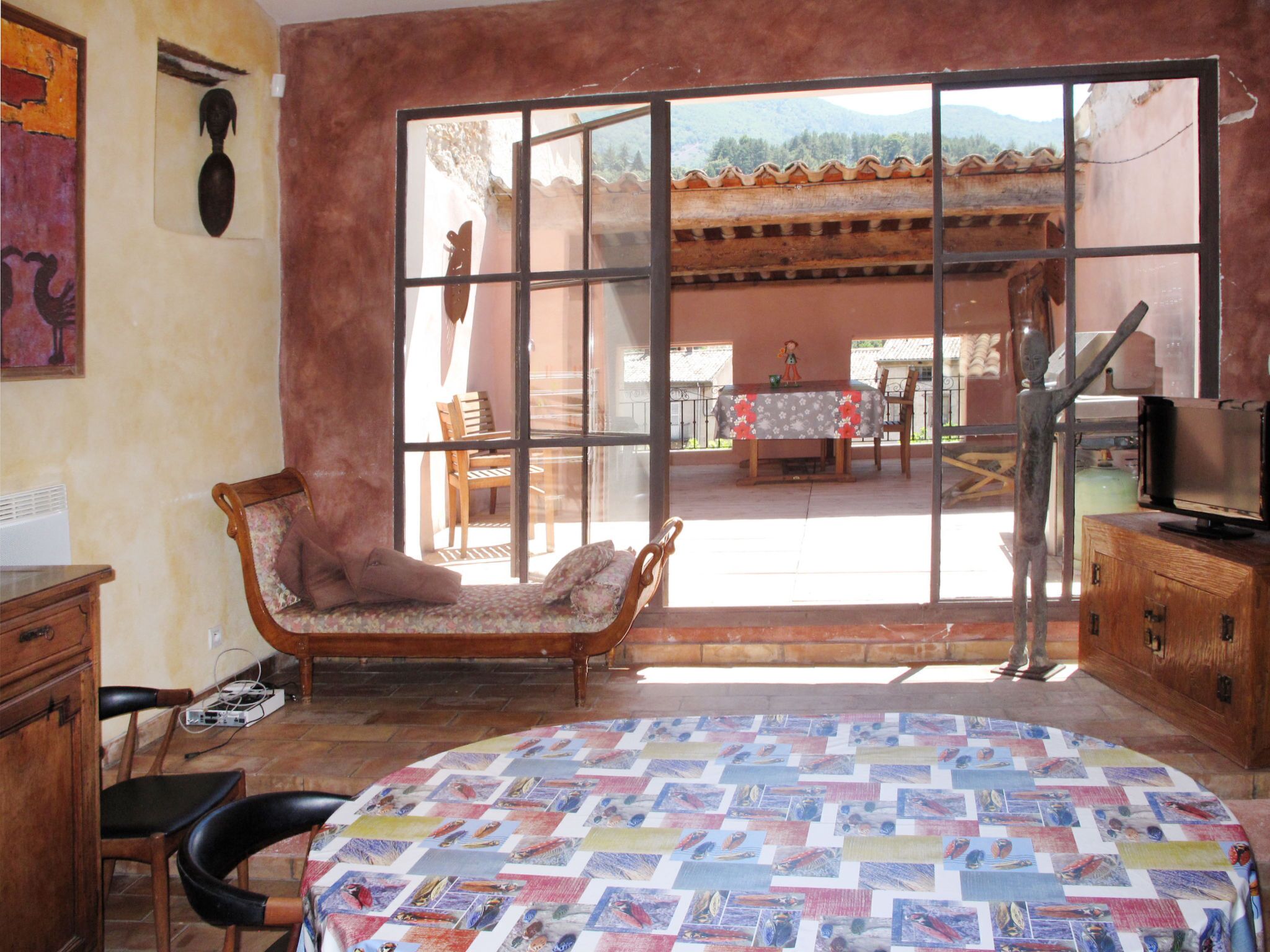 A bedroom with a large window, a bed with a colorful quilt, a wooden chair, a wooden cabinet, and a television.