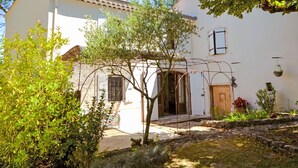 A white building with a tiled roof, a wooden door, and a small garden area with plants and a tree.