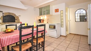 A dining area with a red and white checkered tablecloth, wooden chairs, and a fruit bowl. There is a kitchen with white cabinets, a stove, and a window.