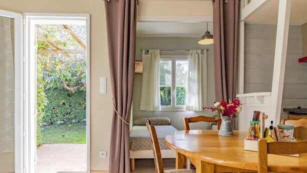 Una habitación acogedora con una mesa de madera, sillas y una cama pequeña. Se ve una ventana con cortinas y vistas a la vegetación.