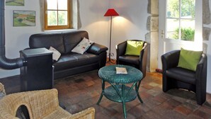 Un salón acogedor con un sofá de cuero negro, un sillón de mimbre verde y una pequeña mesa redonda. Hay dos ventanas con cortinas, una lámpara de pie roja y una pared de piedra.