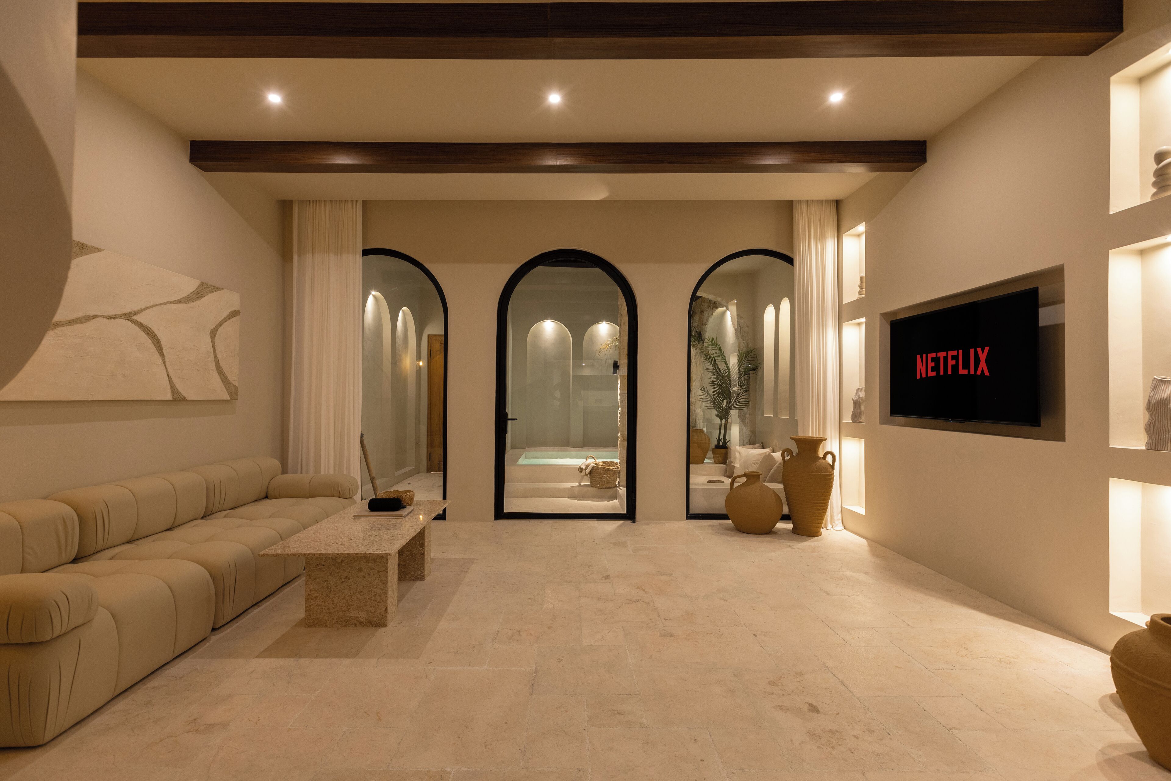 A modern living room with a sofa, a coffee table, and a TV displaying 'NETFLIX'. There are three arched doorways leading to a bathroom.