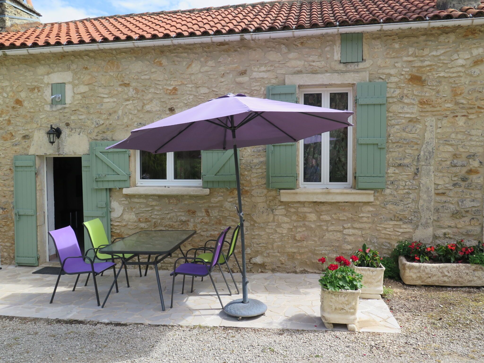 A stone building with a tiled roof, a table with chairs, and a purple umbrella.