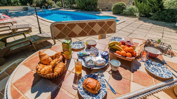 A poolside breakfast setup with croissants, fruits, and beverages on a tiled table. - Le Coustal by Interhome (Blanquefort-sur-Briolance)