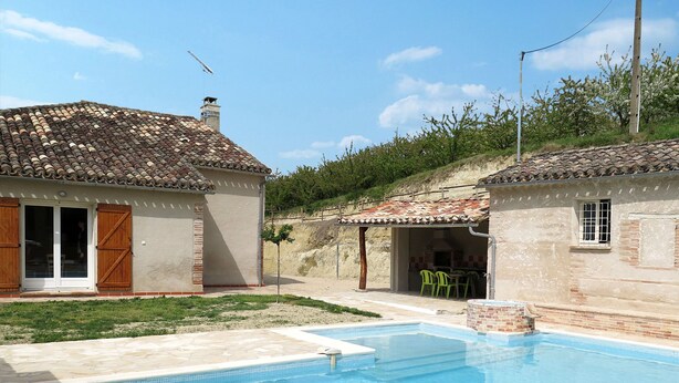 Una piscina con plataforma de baldosas, dos edificios pequeños con tejados de cerámica y una zona ajardinada.