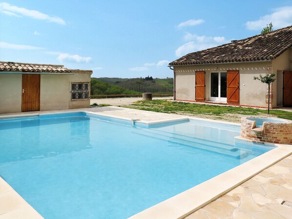 Una piscina rectangular con borde de azulejos, rodeada por una terraza de piedra. Se observan dos edificios con techos de azulejos y puertas de madera, uno de ellos con un pequeño jardín.