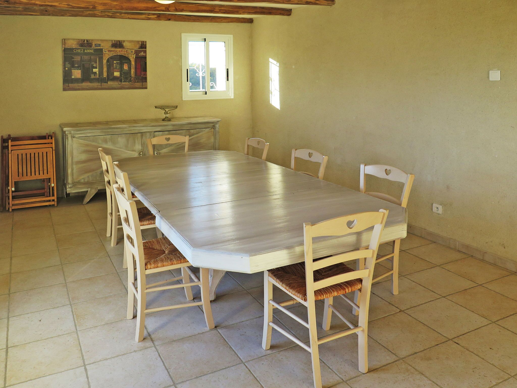 A dining room with a large rectangular table, six wooden chairs with cushioned seats, a small window, and a wall-mounted picture.