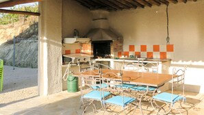 Un patio exterior con suelo de piedra, una mesa de madera, sillas azules, un tanque de gas y un horno de ladrillo.