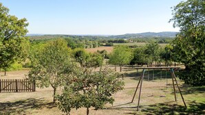A fenced outdoor area with a swing set, trees, and a view of distant hills. - Sarrouil by Interhome (Loubressac)