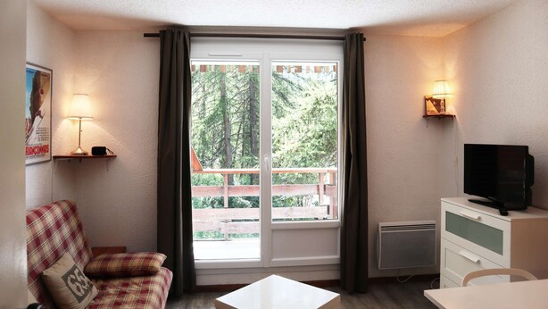 A small, furnished room with a sofa, a television, a table, and a window with curtains. - Fully-equipped studio for 4 people (LES ORRES 1650)